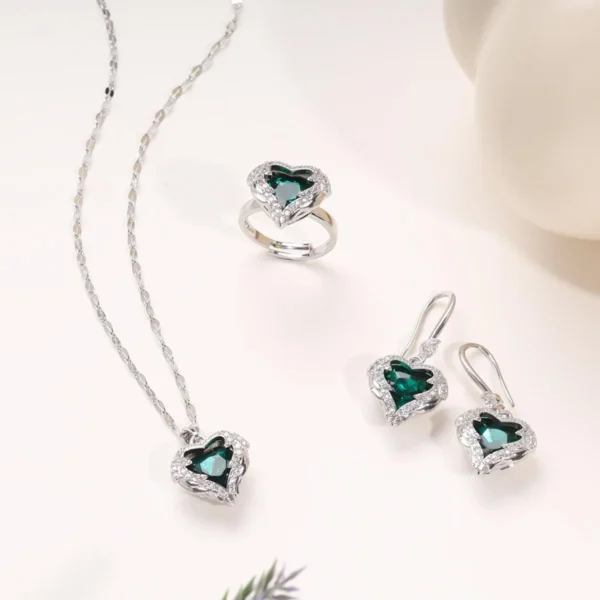 Elegant silver jewelry set featuring a green heart-shaped gemstone necklace, matching earrings, and a ring, all adorned with sparkling white stones, styled on a minimal light background.