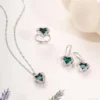 Elegant silver jewelry set featuring a green heart-shaped gemstone necklace, matching earrings, and a ring, all adorned with sparkling white stones, styled on a minimal light background.