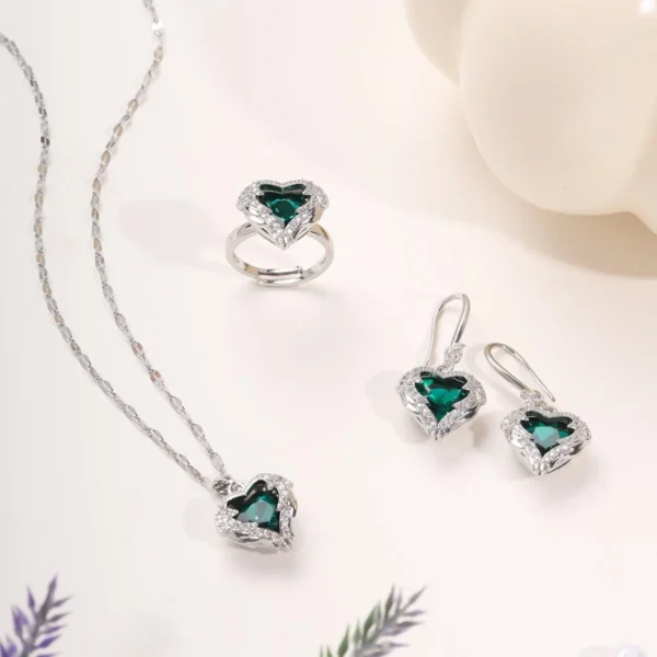 Elegant silver jewelry set featuring a green heart-shaped gemstone necklace, matching earrings, and a ring, all adorned with sparkling white stones, styled on a minimal light background.