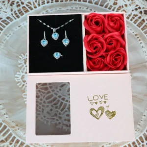 Pink gift box with blue heart-shaped necklace, earrings, and ring set, arranged beside red roses, featuring gold “LOVE” print and clear window.