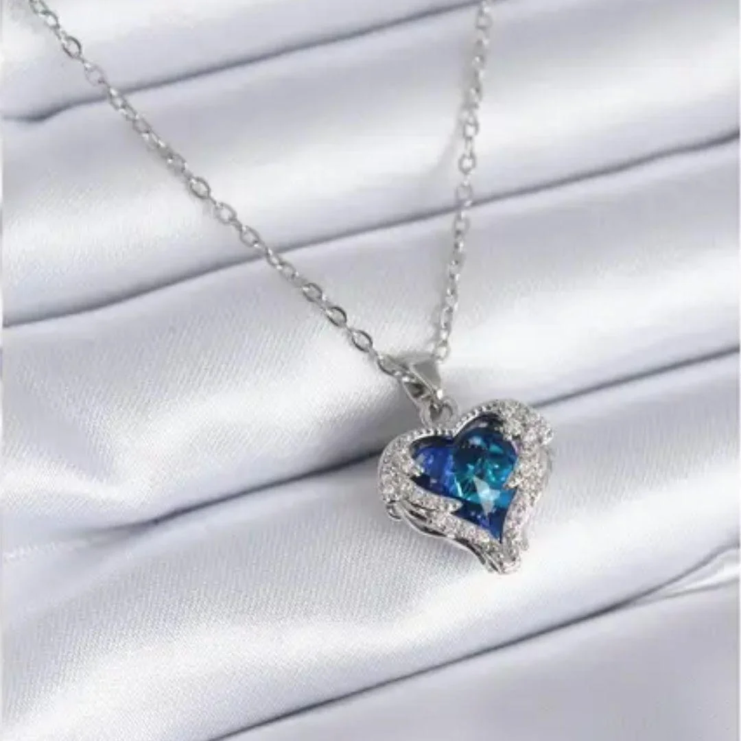Elegant silver chain necklace with a radiant deep blue heart-shaped gemstone pendant, surrounded by shimmering white crystals, resting on a silky white fabric background.