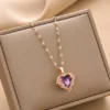 Gold chain necklace with a heart-shaped purple crystal pendant, surrounded by delicate claw-style detailing, displayed with seashells and greenery.