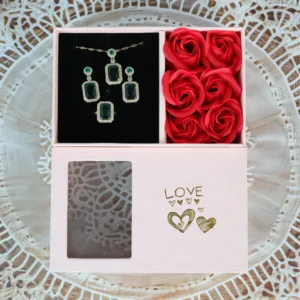 Jewelry gift box with emerald green necklace, earrings, and ring set alongside red artificial roses in a pastel pink “LOVE” box.