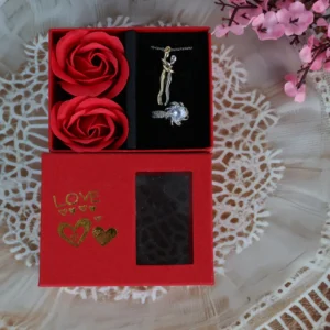 Red “LOVE” gift box with hugging couple necklace, white solitaire ring, and two red roses with transparent display window.