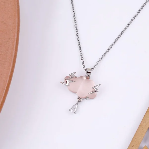 Silver chain necklace with a light pink cloud-shaped pendant, lightning bolt accents, and a teardrop crystal charm, displayed on a terrazzo-style surface.