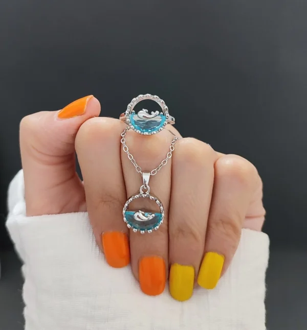 Matching silver necklace and ring set featuring a 3D ocean wave with a dolphin design inside a circular frame, held between fingers with yellow nails.